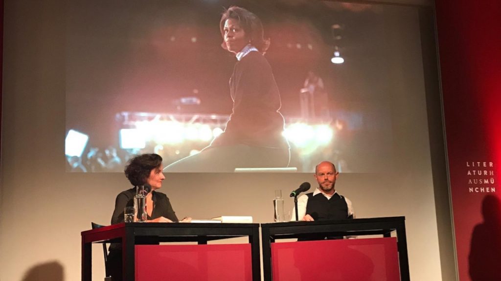 Julia Cortis und Christian Baumann auf der Bühne des Münchner Literaturhauses, im Hintergrund ein übergroßes Portrait von Michelle Obama als Projektion.